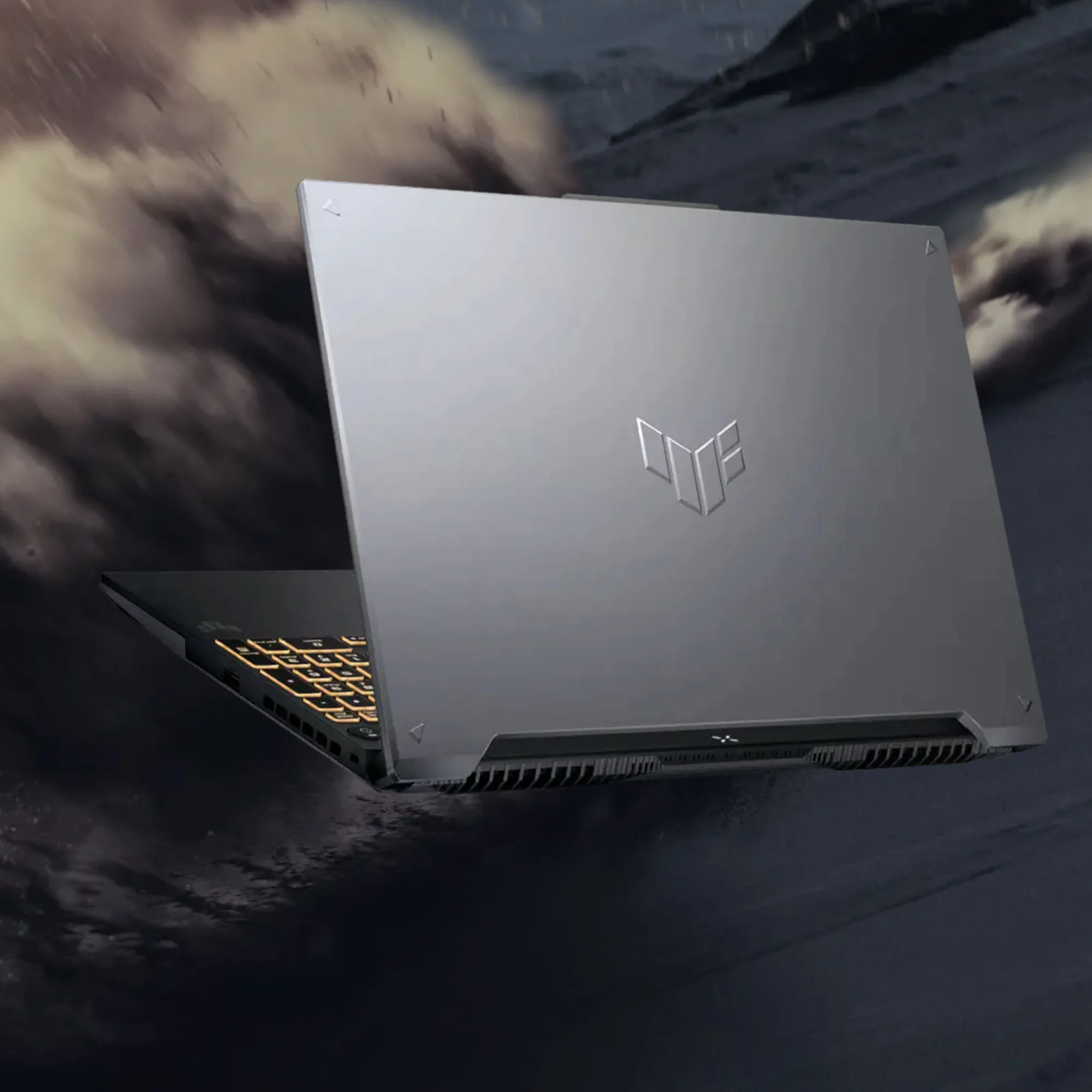 Laptop gamingowy ASUS TUF widziany z tyłu, z podświetlaną na pomarańczowo klawiaturą i charakterystycznym logo TUF na metalicznej pokrywie, na tle dynamicznej scenerii z pyłem i ruchem.