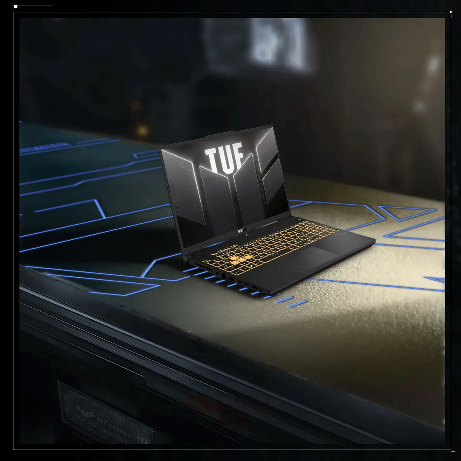 Laptop ASUS TUF z podświetlaną na żółto klawiaturą i ekranem z dużym logo TUF, ustawiony na futurystycznym blacie z niebieskimi liniami LED.