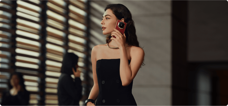Kobieta w eleganckiej czarnej sukni trzyma w ręku bordowy smartfon Huawei Mate X7, w tle dwie rozmyte postaci