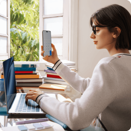 Siedząca przy biurku i otwartym oknie kobieta w okularach pracuje na laptopie trzymając w dłoni smartfona Huawei Nova 14 pro