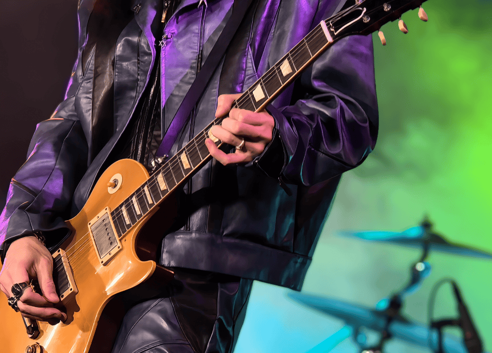 Muzyk w skórzanej kurtce i spodniach gra na gitarze elektrycznej, za nim talerze zestawu perkusyjnego