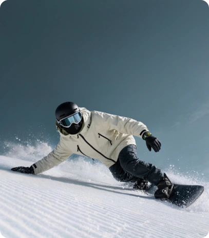 Snowboardzista, w jasnej kurtce i czarnym kasku sunie dynamicznie po zaśnieżonym stoku, przytrzymując się dłonią podłoża