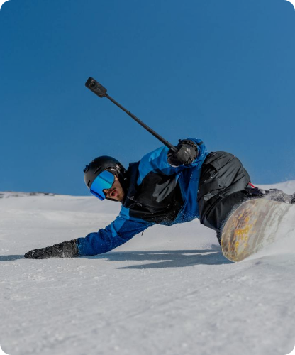 Snowboardzista w niebiesko-czarnej odzieży sportowej i kasku z goglami, wykonujący dynamiczny skręt na śnieżnym stoku, trzymający kijek z kamerą sportową.