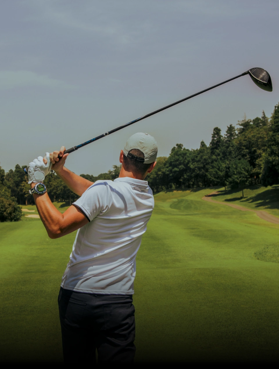 Golfista ubrany w białą koszulkę i czapkę wykonujący zamach kijem golfowym na zielonym polu golfowym, z widocznymi drzewami w tle.