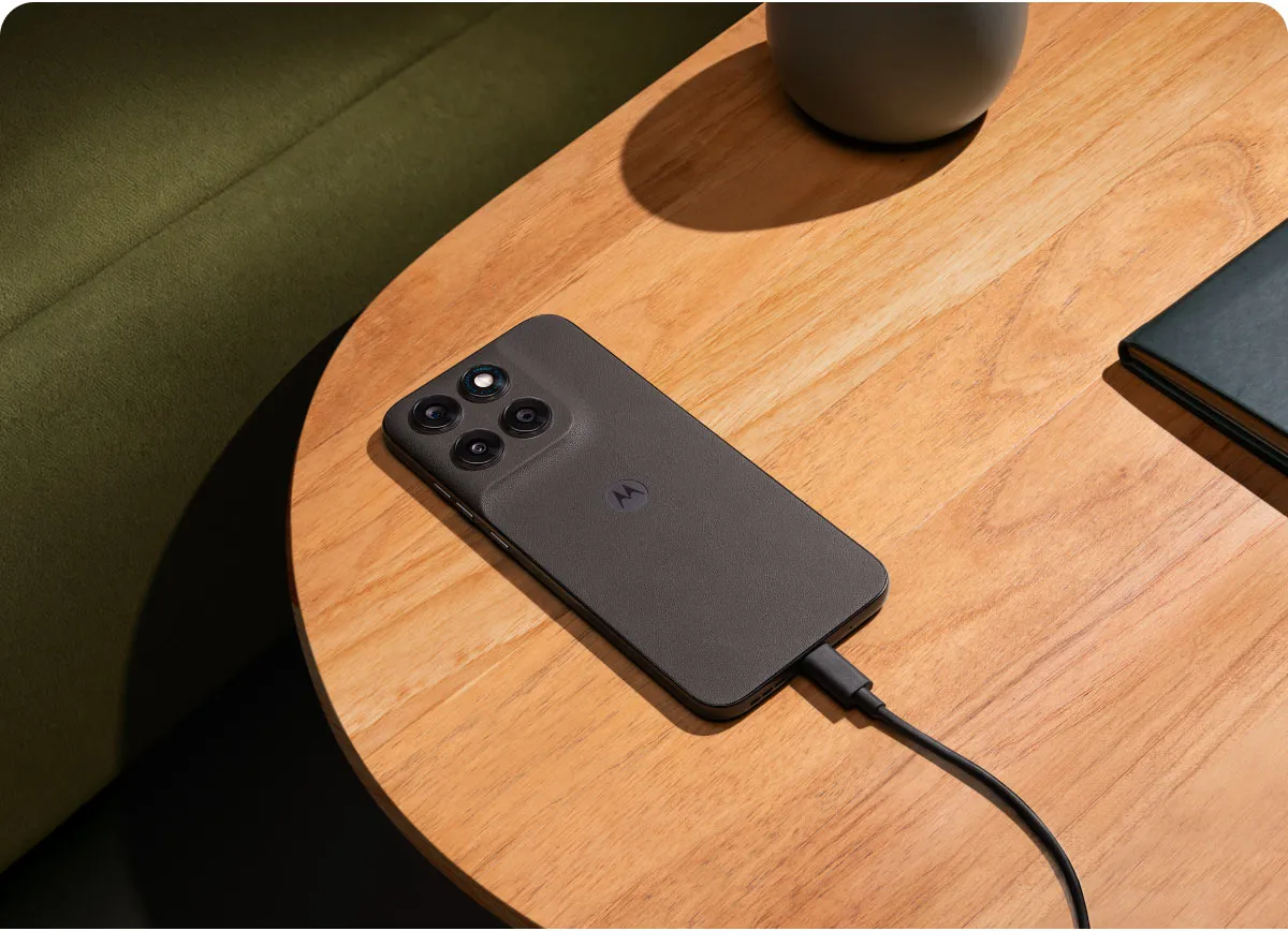 Smartfon moto g77 w ciemnym kolorze leżący na drewnianym stoliku kawowym, podłączony kablem do ładowarki