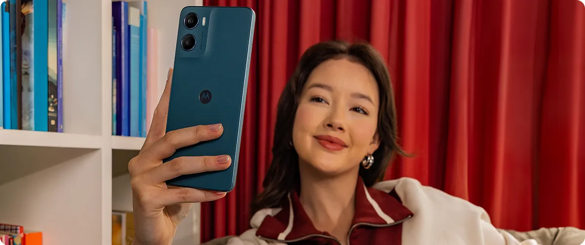 Kobieta robiąca selfie niebieskim smartfonem Motorola moto g06 POWER na tle półki z książkami