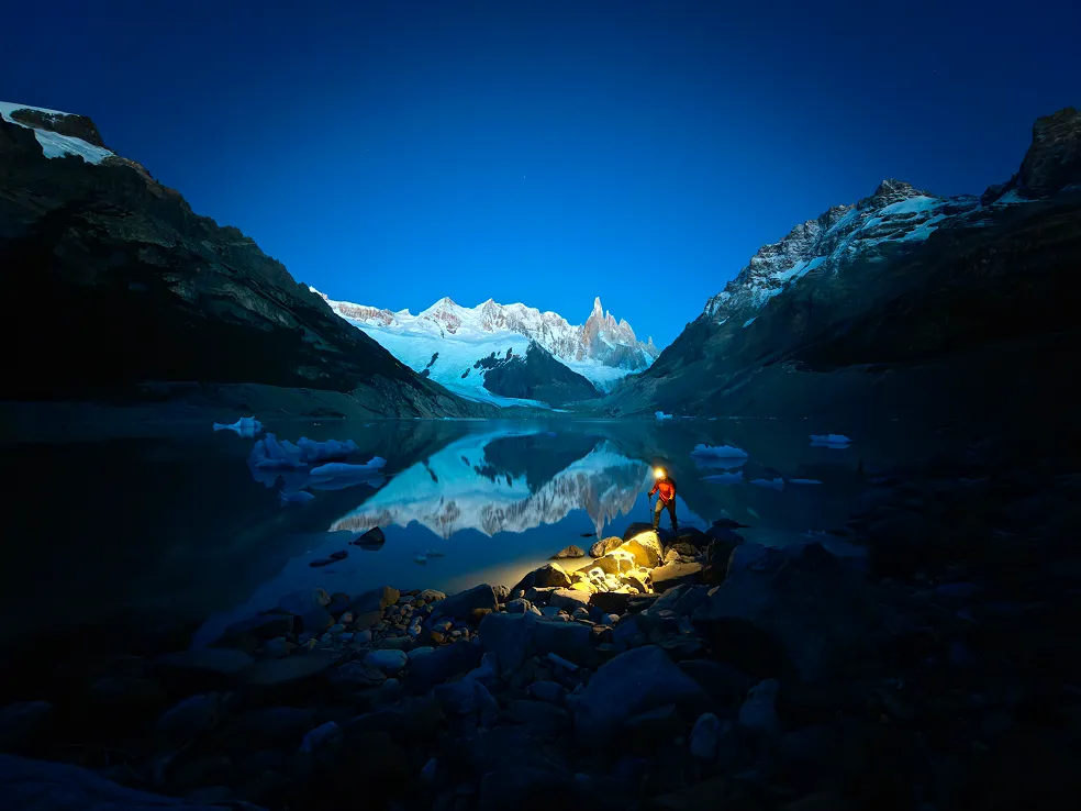 Nocne zdjęcie górskiego jeziora z odbiciem szczytów i sylwetką alpinisty z latarką