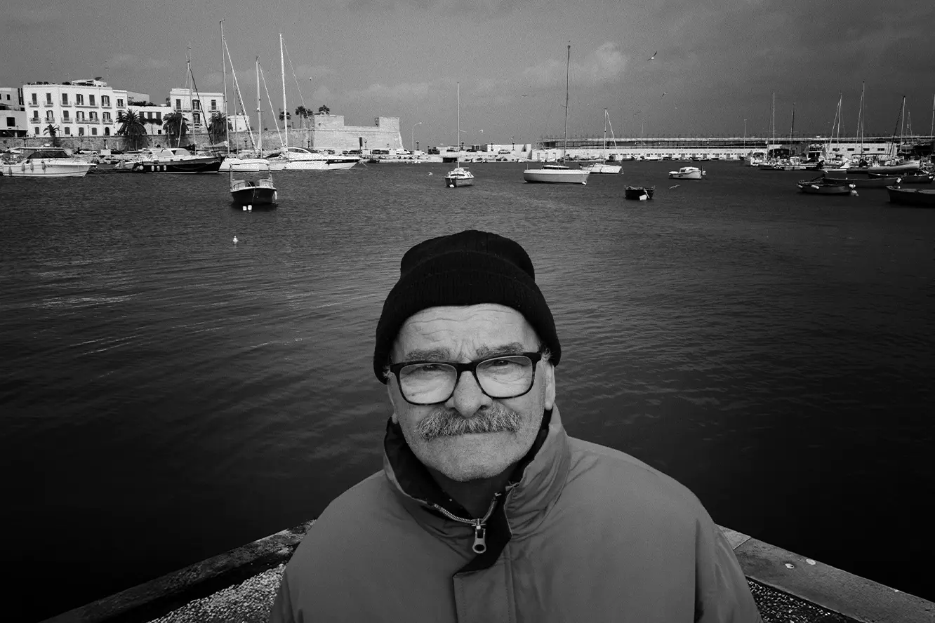 Portret starszego mężczyzny w czapce i okularach nad wodą, w tle port z łodziami i zabudową