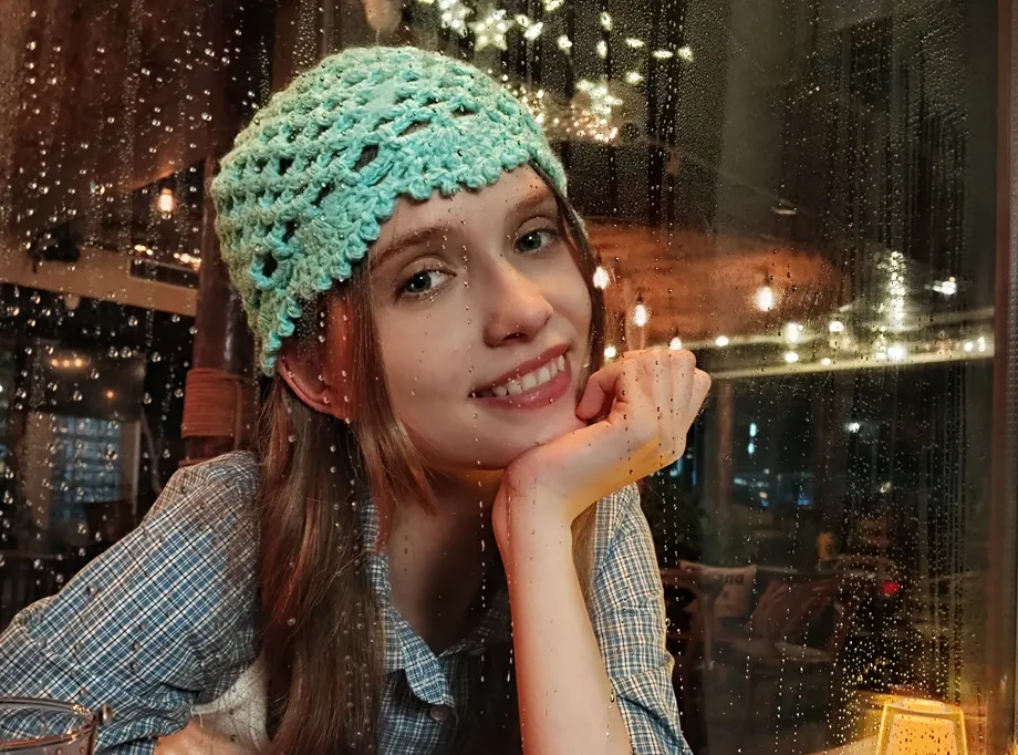 Portret uśmiechniętej kobiety w zielonej czapce, wykonany wieczorem przez szybę z kroplami deszczu