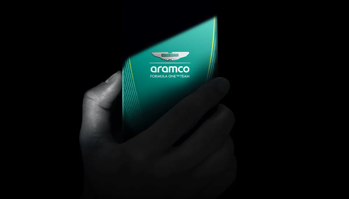 Dłoń trzymająca smartfon z widocznym srebrnym logo Aston Martin na zielonym tle.